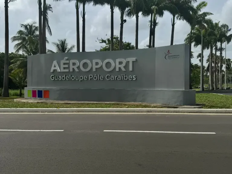 La photo montre le panneau d'entrée de l'Aéroport Guadeloupe Pôle Caraïbes. Le panneau est grand et rectangulaire, posé au niveau du sol devant une rangée de grands palmiers. Le texte "AÉROPORT" est affiché en grosses lettres blanches sur la partie supérieure du panneau, et en dessous, en plus petites lettres, il est écrit "Guadeloupe Pôle Caraïbes". Le fond du panneau est gris, et il y a un logo à droite qui semble être celui de l'aéroport, combinant probablement des éléments symboliques de la Guadeloupe. Le panneau est situé juste à côté d'une route, visible par les véhicules qui passent.