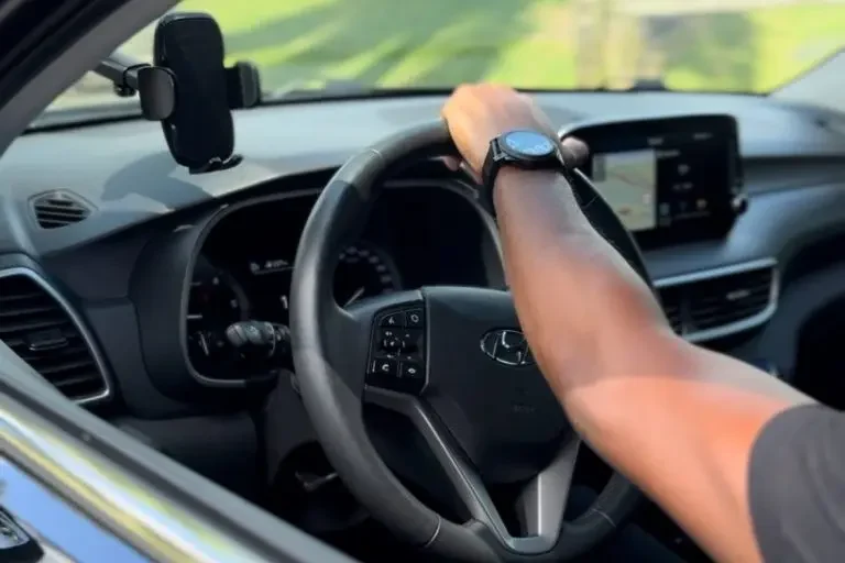 Cette image montre l'intérieur d'un véhicule, capturant la vue depuis le siège passager avant, tourné vers le conducteur. Le conducteur, probablement un chauffeur privé en Guadeloupe, tient fermement le volant avec sa main gauche, reflétant une posture professionnelle et attentive. Le tableau de bord est moderne avec un écran tactile affichant des informations de navigation, ce qui est typique des véhicules utilisés par les services de chauffeur privé pour offrir une expérience de conduite efficace et confortable. Le cadre interne est propre et bien entretenu, suggérant un service de haute qualité pour les passagers recherchant des transports privés en Guadeloupe.